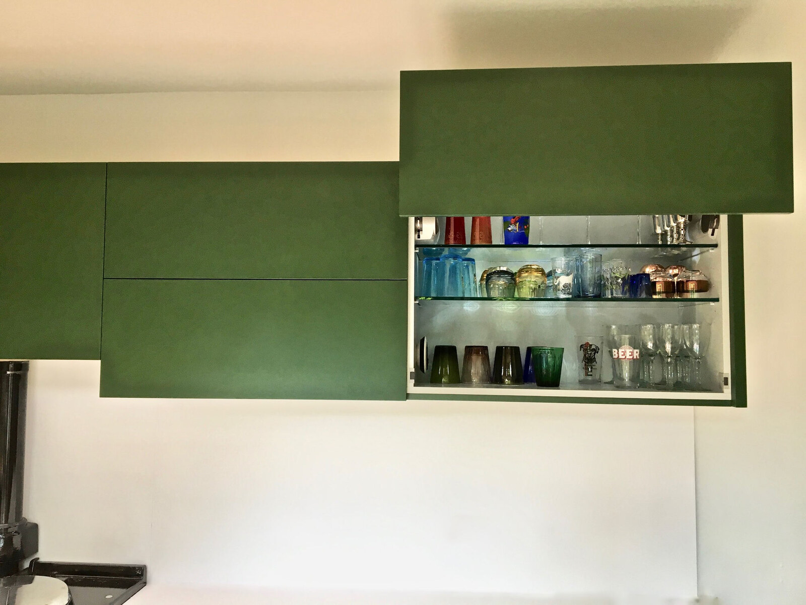 Green handleless wall units, one open showing colourful glassware on glass shelves
